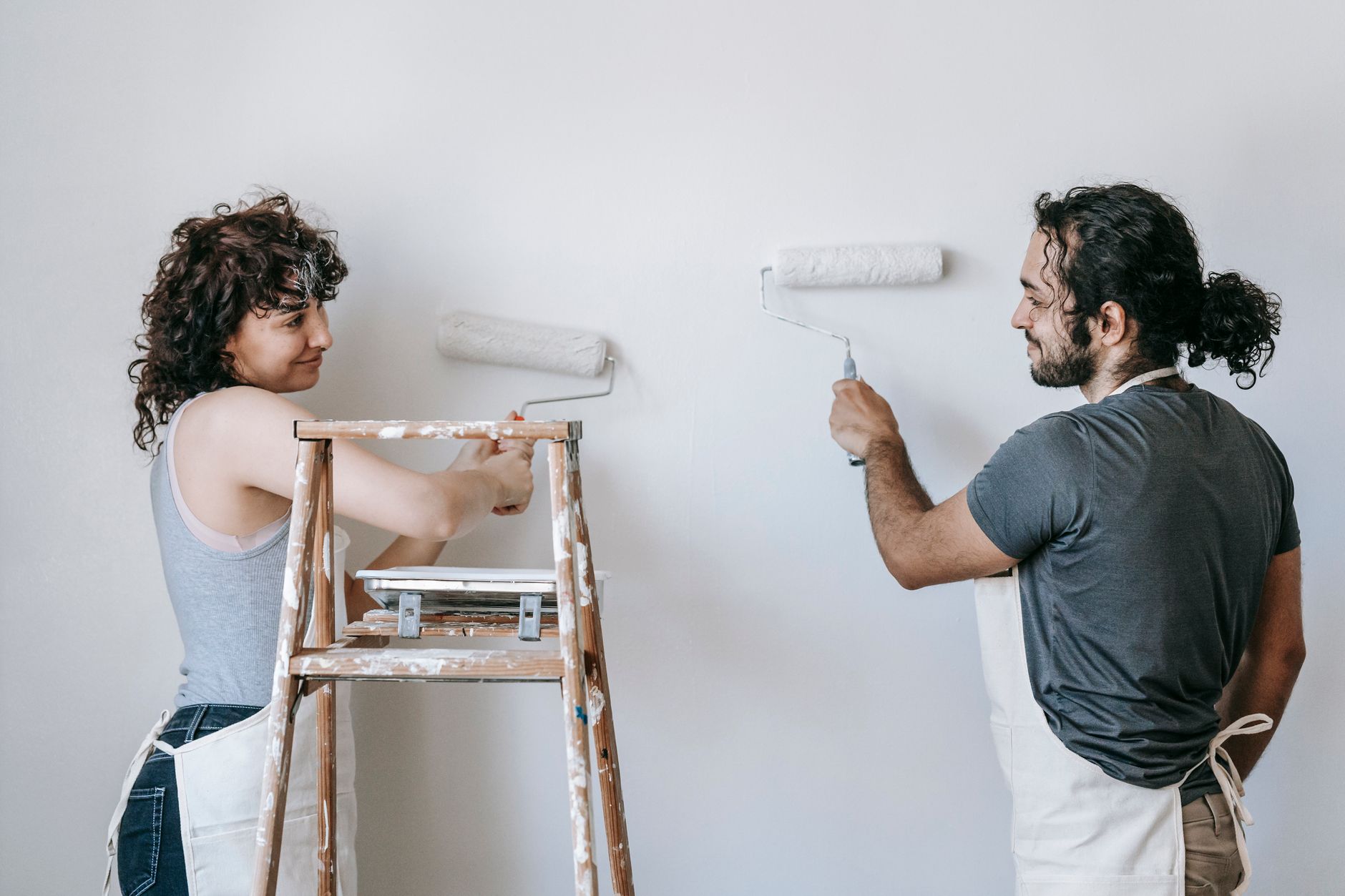 Cheerful adult ethnic couple in aprons looking at each other while painting wall with rollers during in Santa Monica, CA